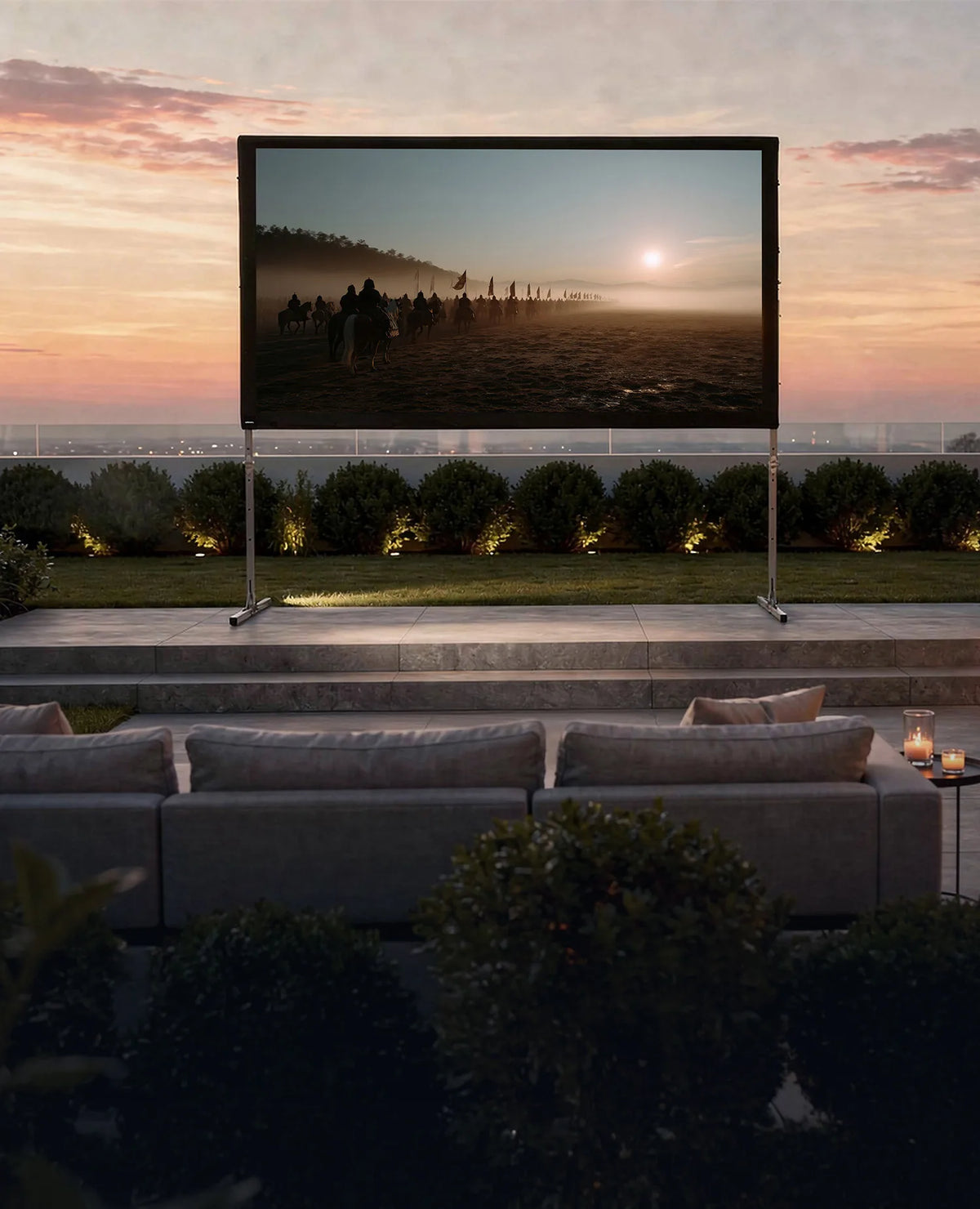 Luxurious outdoor home cinema in the evening: in a modernly designed garden, a celexon Folding Frame Projector Screen Mobile Expert with rear projection is set up on a terrace, displaying an atmospheric film scene featuring a rider at sunrise. In front of it, elegant outdoor sofas with cushions and candles invite relaxed viewing. On the right, a stylish illuminated outdoor bar with bar stools, glasses, and bottles complements the scene.