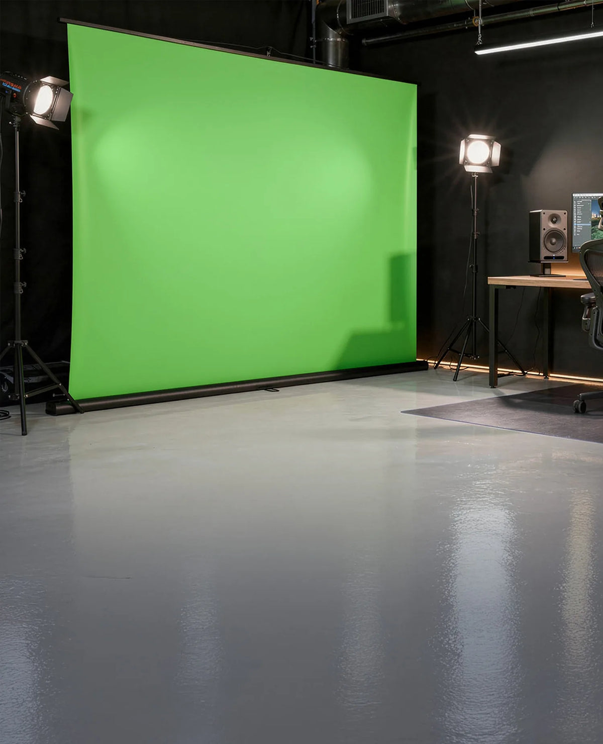 A professionally designed studio and work environment, with a large, free-standing celexon Mobile Lite Chroma Key green screen positioned on the left, while on the right there is a modern workstation featuring a minimalist wooden desk, two monitors displaying video editing software, and two speakers.
