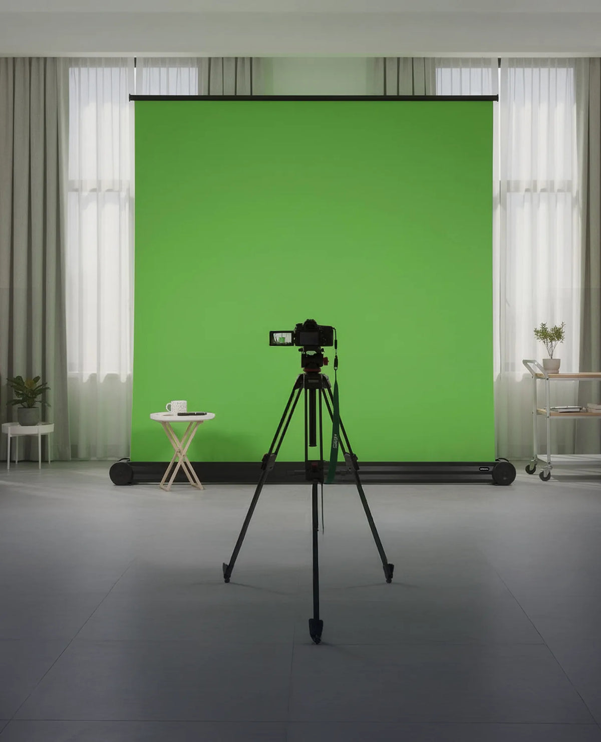 Professional studio setup for video or photo production. In the center of the room stands a large, free-standing celexon Mobile Chroma Key green screen. Positioned centrally in front of it is a camera mounted on a tripod.