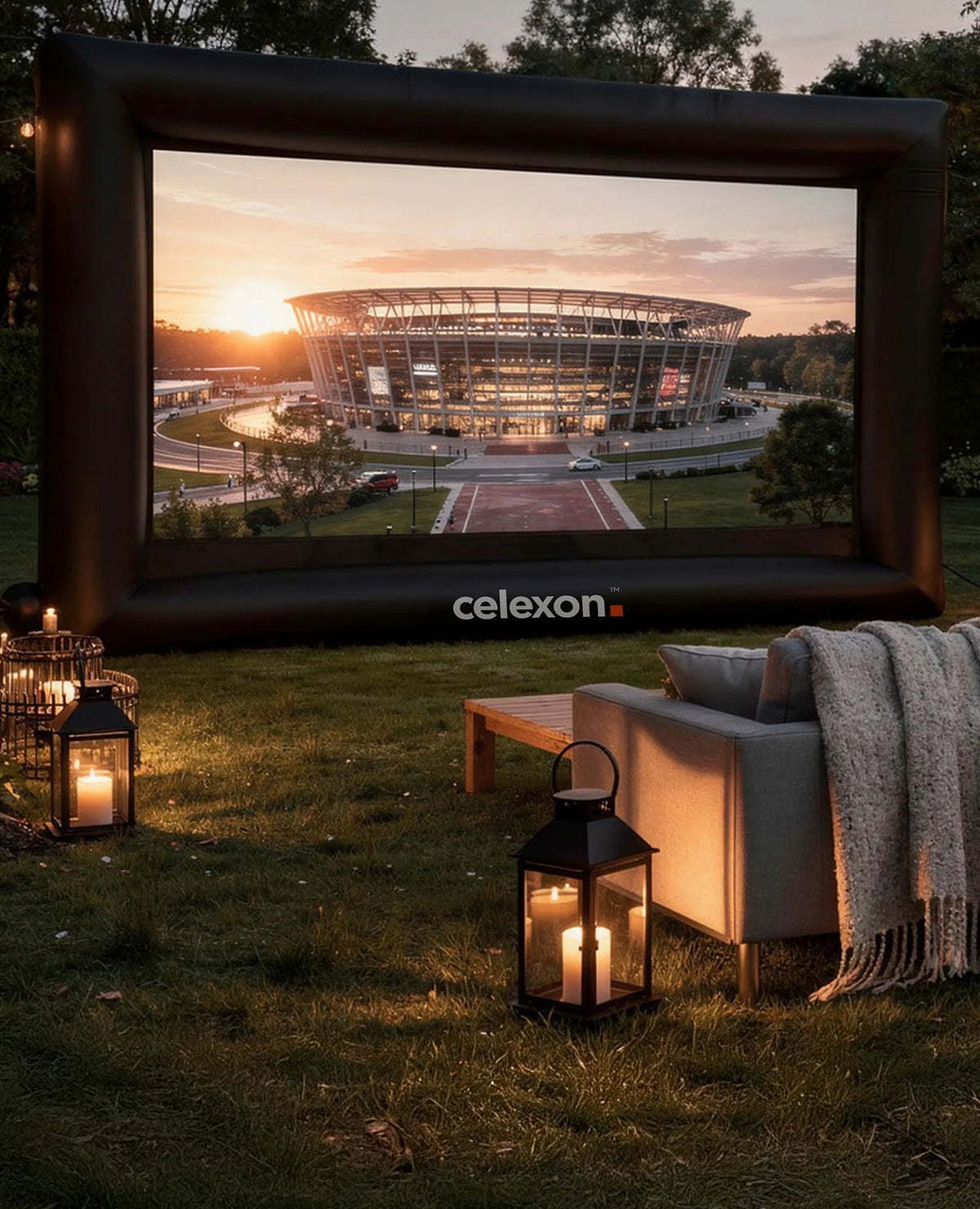 Atmospherically staged outdoor home cinema in a garden at dusk. At the center is the celexon inflatable outdoor projector screen INF200, displaying a modern stadium bathed in the warm light of the sunset. In front of it, cozy sofas with blankets invite people to sit, while lanterns with candles and string lights create a homely atmosphere.