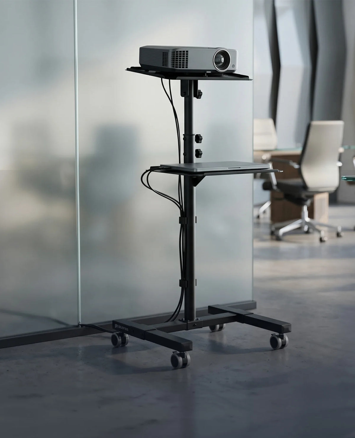 A mobile celexon PT3020 projector trolley with castors and height-adjustable shelves holding a projector and a laptop. The trolley is positioned in a modern office environment with glass walls and a meeting room in the background.