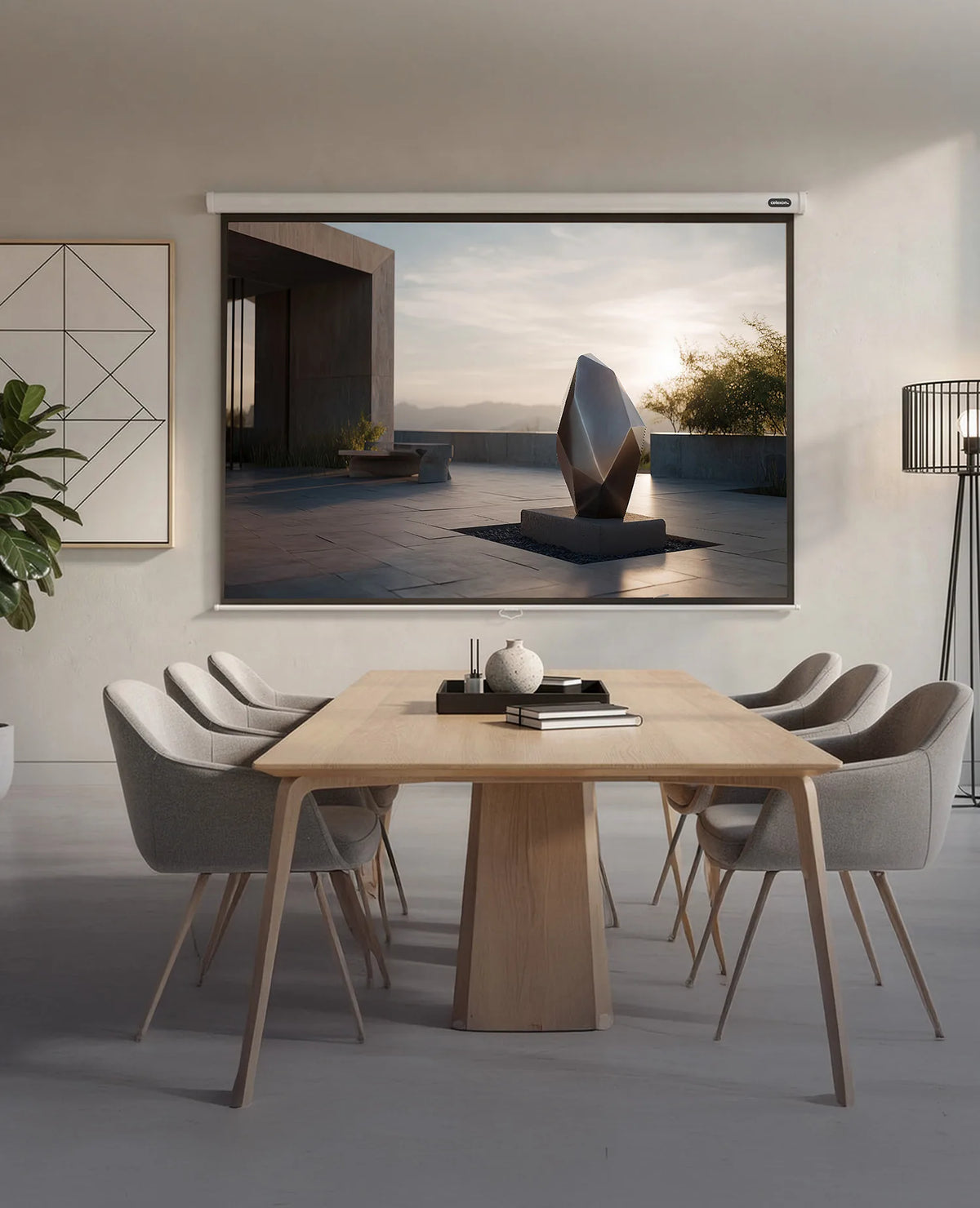 The image shows a modern meeting room with a wooden table and upholstered chairs, in which a celexon Manual Economy projector screen displays a calm outdoor architectural scene featuring an abstract sculpture in warm evening light.