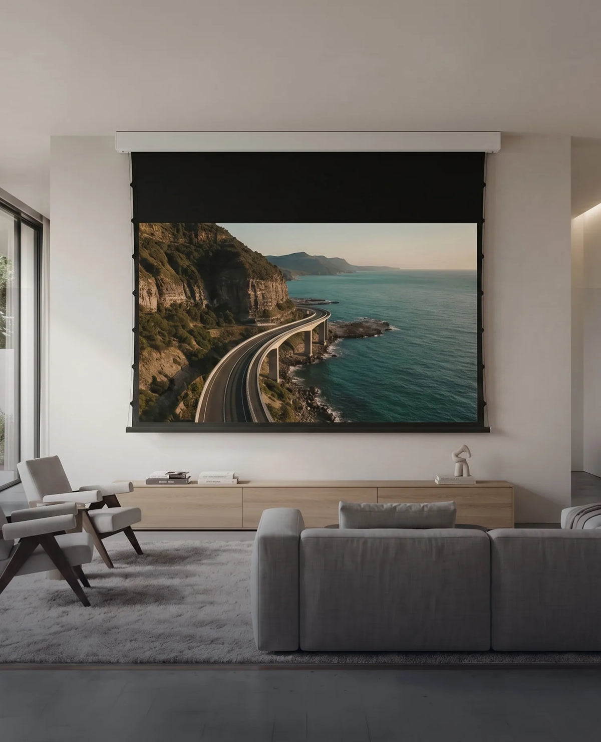 A modern living room featuring a celexon SOUNDVISION Premium Plus Screen Electric Tension, displaying a coastal road along the sea. Light-colored seating, a low sideboard, and minimalist furnishings create an elegant home cinema atmosphere.