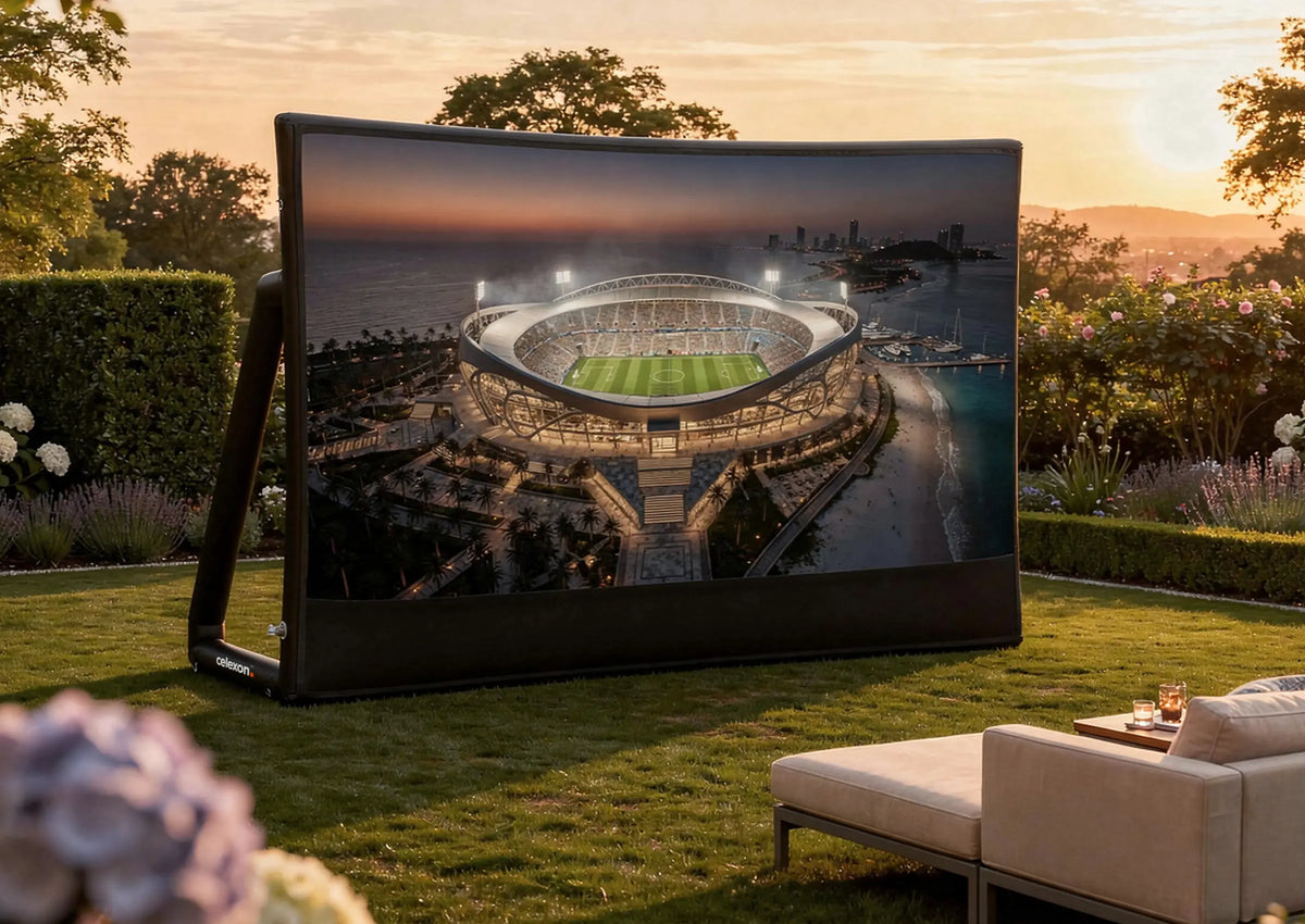 A celexon inflatable outdoor projection screen Economy INF in a garden at sunset; on it, a brightly lit football stadium by the coast can be seen. In the foreground, there is a lounge chair and small table decorations, surrounded by a well-kept lawn, flowers, and trees, creating a relaxed open-air cinema atmosphere.