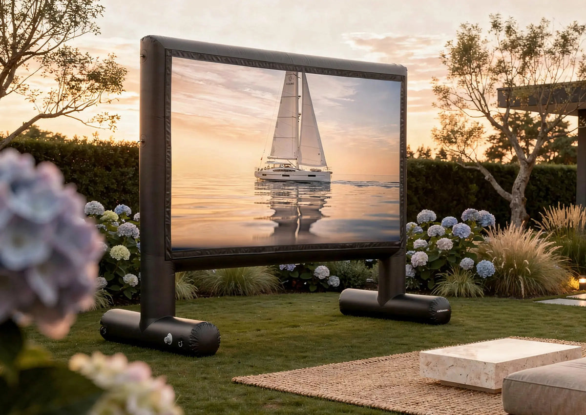 An celexon inflatable outdoor projection screen Professional INF in a well-kept garden at sunset; on it, a sailboat can be seen on calm water. Surrounded by flowers, shrubs, and modern garden furniture, the scene feels like a cozy open-air cinema.