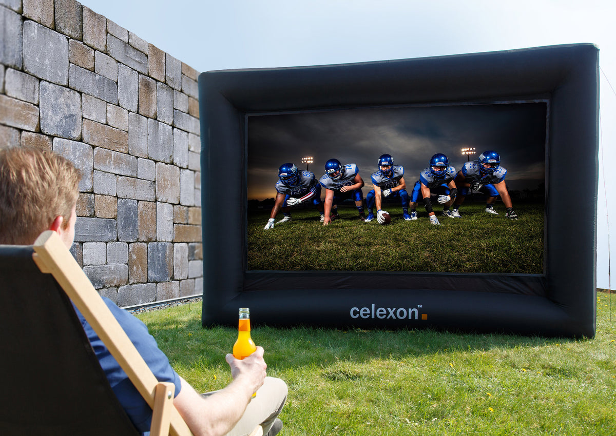 Outdoor scene with a large, inflatable celexon inflatable outdoor projector screen INF200 set up on a grassy area in front of a tall stone wall. An American football game is projected onto the screen, showing several players in full gear standing in a starting formation on the field. In the foreground on the left, a person sits relaxed in a deck chair, looking toward the screen and holding a beverage bottle in their hand.